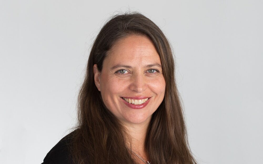 En leende, brunnet kvinna som sitter med kroppen lutad åt höger och ansiktet rakt mot kameran.