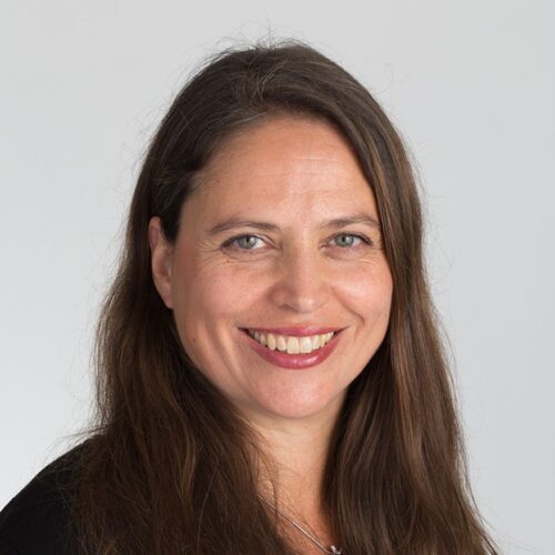 En leende, brunnet kvinna som sitter med kroppen lutad åt höger och ansiktet rakt mot kameran.